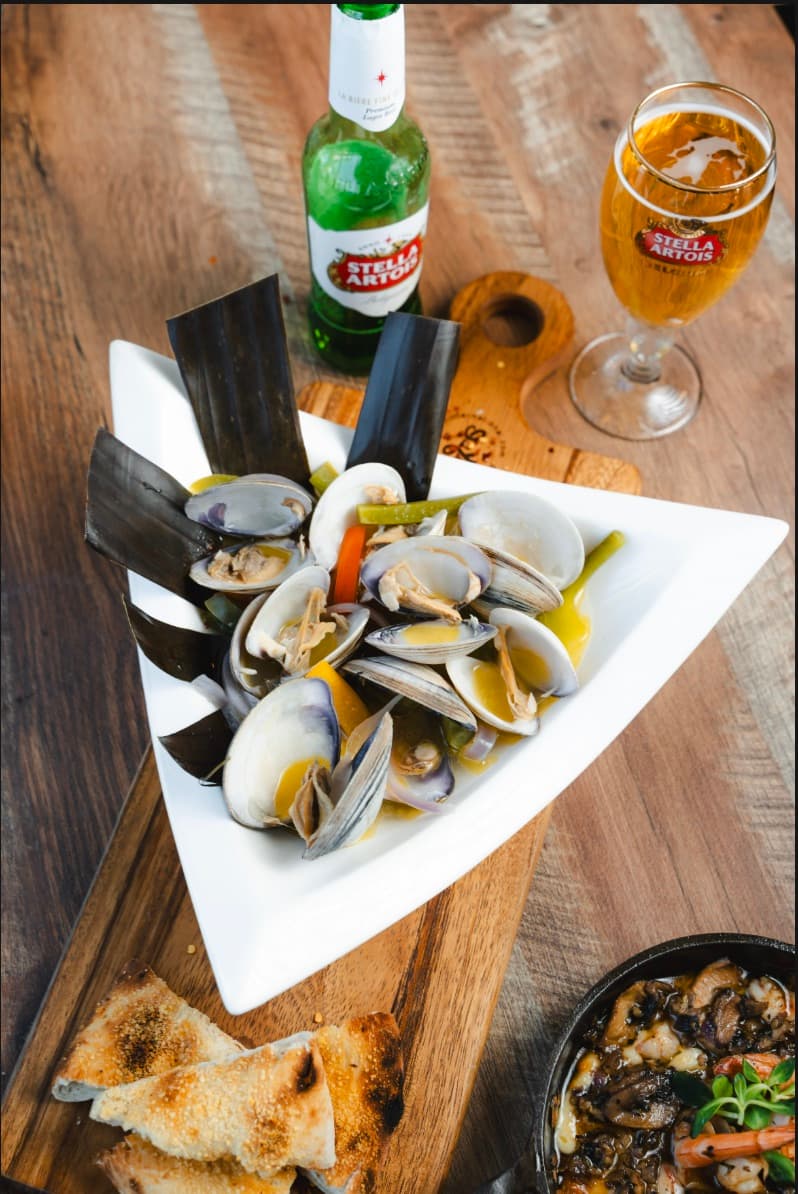 Almejas al vapor en caldillo, servidas en plato triangular con pan tostado y una cerveza Stella Artois.