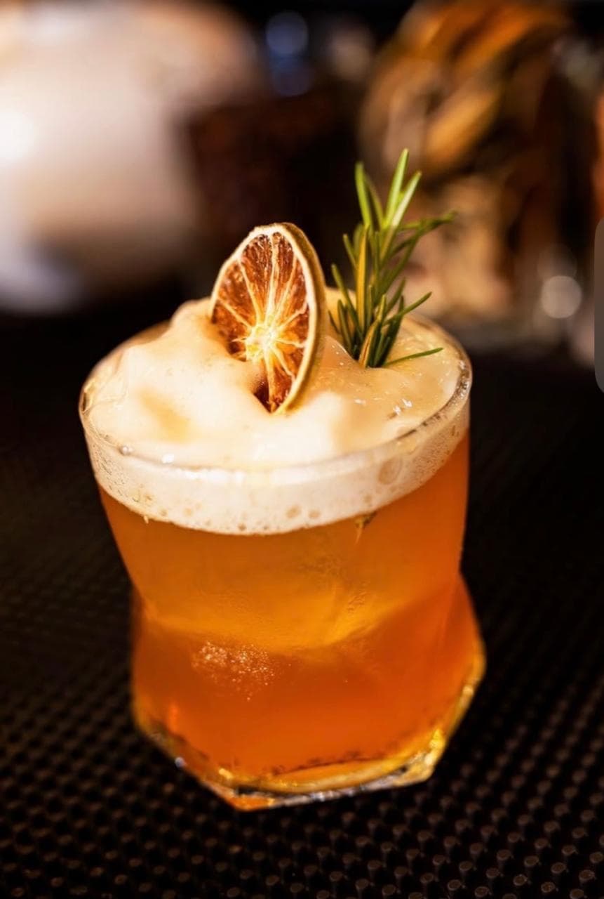 Coctel ámbar con espuma, decorado con una rodaja de cítrico deshidratado y una ramita de romero.