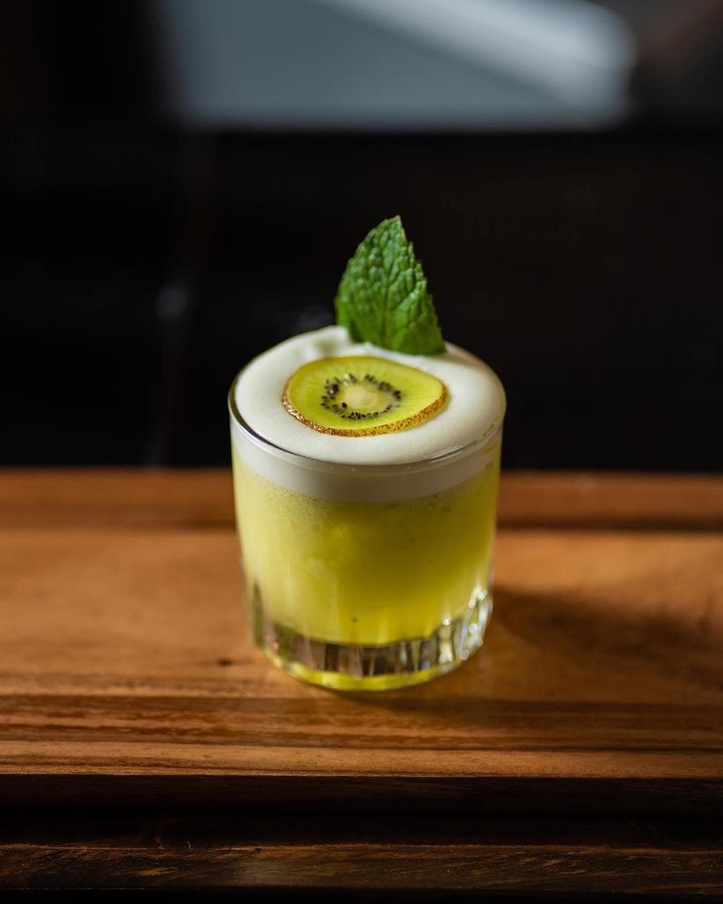 Coctel verde con espuma y una rodaja de kiwi sobre una mesa de madera.