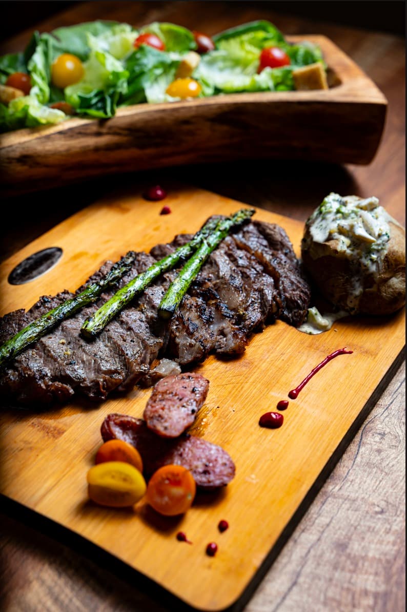 Rib Eye a la brasa sobre tabla de madera, con espárragos asados, papa al horno, rodajas de chorizo argentino y tomates cherry, junto a una ensalada.
