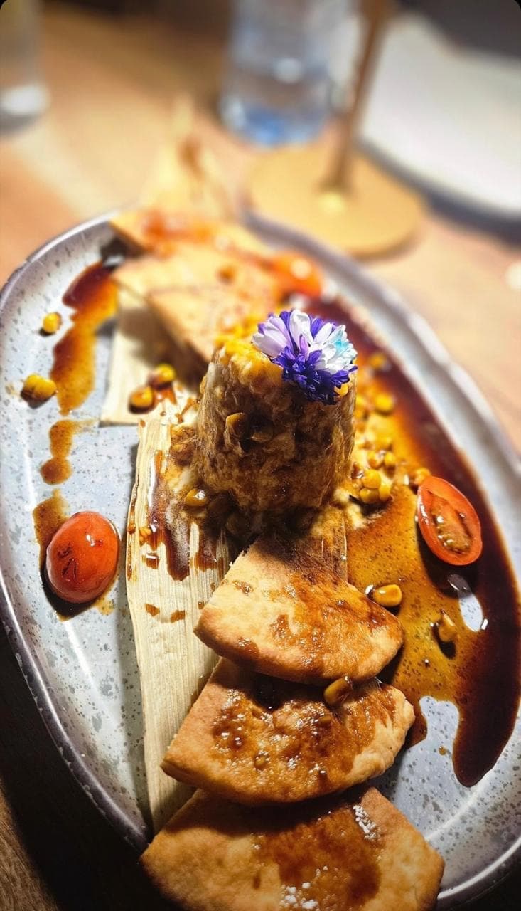 Tamal con costra crujiente sobre hoja de maíz, bañado en salsa de achiote, acompañado de totopos y flor comestible.